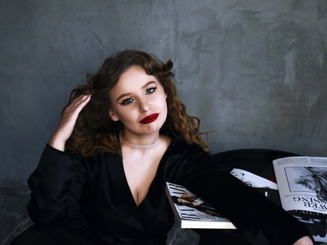 KyleJackson with curly brown hair, red lipstick, black V-neck dress, silver necklace, sitting, hand in hair, magazines on black table, gray concrete wall background.