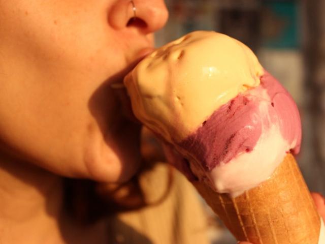 JaneRoa's face, nose ring, and lips are around a double-scoop ice cream with yellow, pink, and white flavors. Her hand holds the waffle cone.