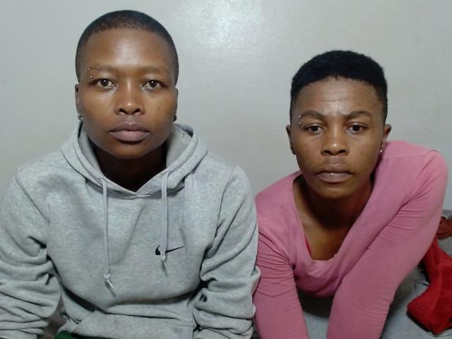 Two sexyboobaex5s sitting; left one in gray Nike hoodie, right one in pink long-sleeve shirt, both with short black hair, looking serious, against plain background.