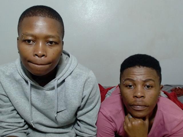 Two sexyboobaex5s with short black hair, one in a gray hoodie, the other in a pink shirt, both with nose piercings, sitting closely, looking at the camera.