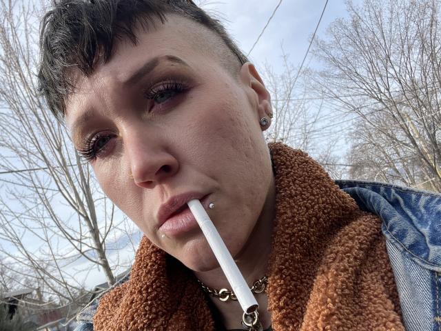 Scarlette, espiègle, avec ses courts cheveux noirs, ses yeux marrons et son piercing au nez, tenant une cigarette dans sa bouche, portant une veste marron duveteuse et une chaîne en or..
