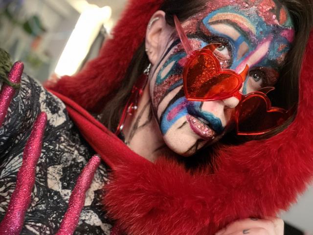 NaughtyScarlette, avec du maquillage coloré sur le visage, des lunettes en forme de cœur rouges et une capuche en fourrure rouge, portant un haut noir et rose à motifs, regardant l'appareil photo avec un sourire espiègle..