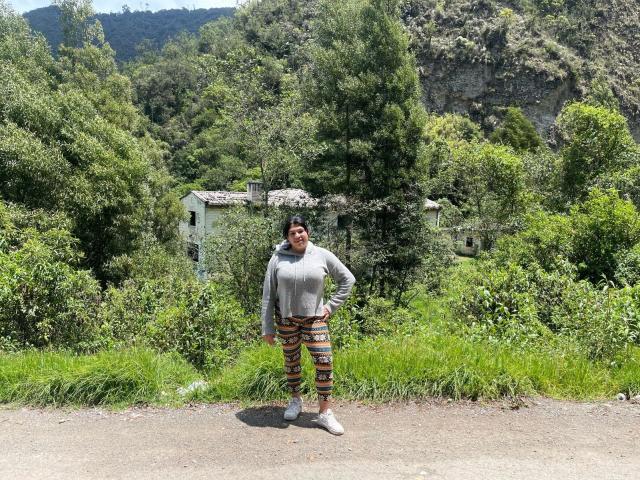 AlmaWhite69 stands in a dirt path wearing a grey hoodie and colorful, patterned leggings with white sneakers, surrounded by lush green trees and a rustic, partially hidden house.