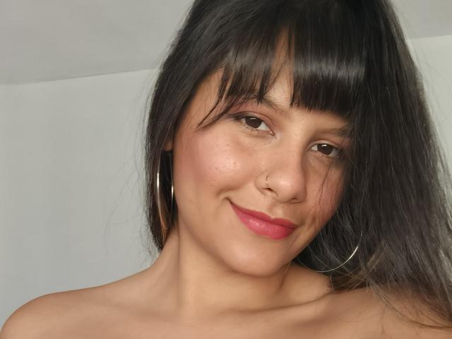 HottPam69 with dark brown hair, bangs, and hoop earrings, smiling with pink lipstick, nose ring, and bare shoulders, showing cute, freckled skin and a slight blush.