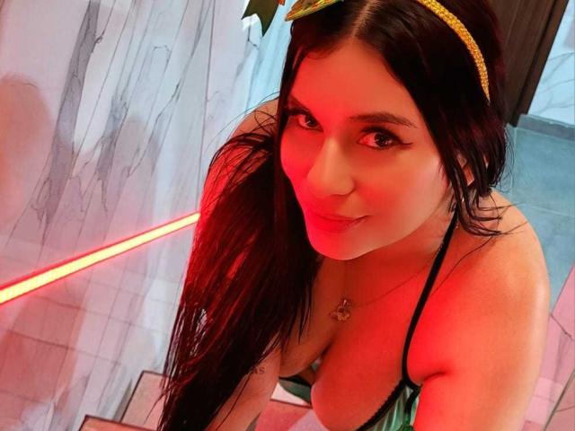 AliceNova with long dark hair, wearing a gold headband and green bra, showing off her tits and smiling with red lighting. She has a heart necklace and eyeliner.