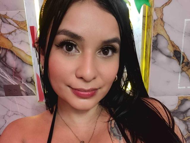 AliceNova, con i capelli neri, gli occhi marroni e il rossetto rosa, sorride alla fotocamera. Indossa un top nero con spalline e un piccolo orecchino a cerchio sul lato destro..
