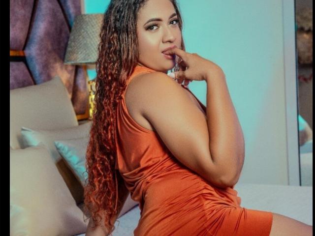 Alejandrawalker03 in an orange dress, sitting on a bed, looking back, finger in mouth, curly red-orange hair, tan skin, showing ass, bedroom setting, seductive expression.