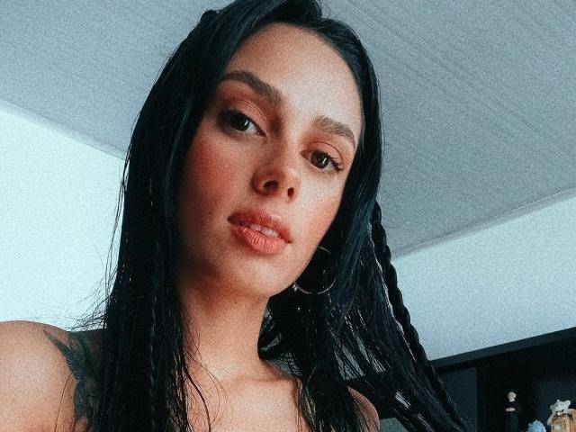 AnaisHilton has wet black hair in braids, hoop earrings, and a tattoo on her right shoulder. She's looking down with slightly parted lips and brown eyes.