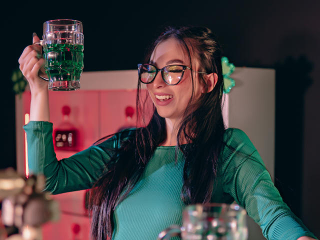 CamilleRios with glasses, green ribbed shirt, holding a glass of green liquid, smiling, dark hair, medium tits, raised arm, dim lighting, semi-sheer green scarf in background.