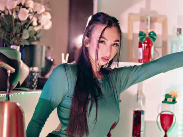 CamilleRios with long dark hair, green ribbed top, arm raised, seductive gaze, pouty lips, small nose, slight makeup, indoors, red and green decor, soft lighting, sexy.