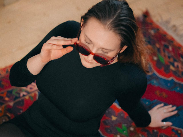 MaryWaddell with red sunglasses, black ribbed long-sleeve top, and brown hair, holding glasses, looking down, seated on a colorful patterned rug, arms slightly spread.