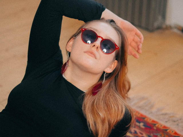 MaryWaddell with red sunglasses, black long-sleeve shirt, and arm raised, showing off her tits. She's lying on a rug with wooden floors and a radiator in the background.