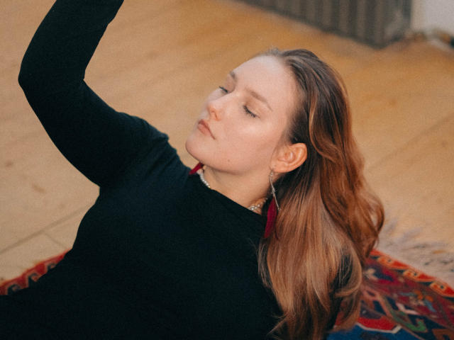 MaryWaddell with her arm raised, wearing a black sweater, red earrings, and a necklace, lying on a colorful rug, eyes closed, wavy brown hair, soft light.