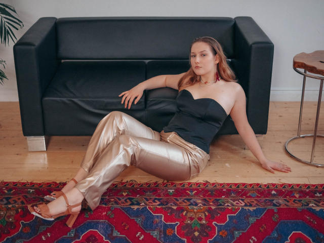 MaryWaddell with small tits, sitting on wooden floor, wearing black strapless top, shiny gold pants, brown sandals, red earrings, black couch, wooden table, red patterned rug, relaxed.