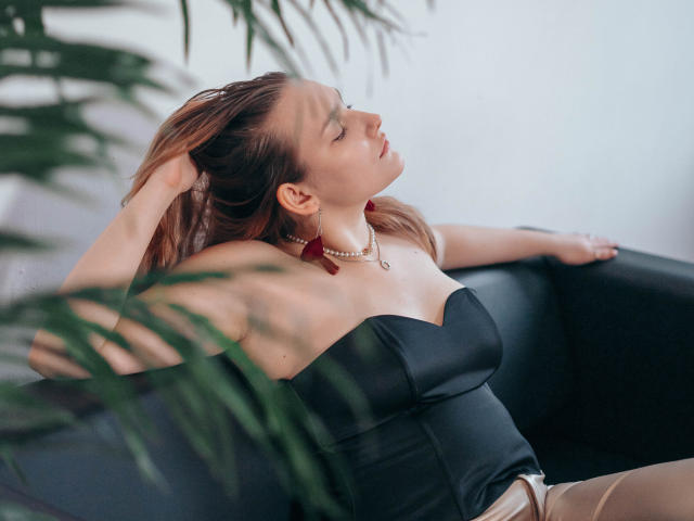 MaryWaddell with her tits in a black strapless top, arms raised, eyes closed, red earrings, pearl necklace, leaning on a black couch, brown hair, potted plant in foreground.