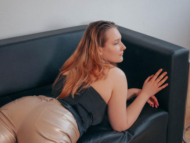 MaryWaddell is lying on a black leather couch, her ass up, wearing shiny gold pants and a black strapless top, with blonde hair and a side profile.