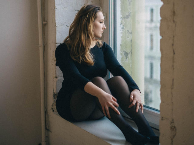 MaryWaddell with wavy brown hair, black long-sleeve dress, and black tights, sitting on a windowsill, legs crossed, hands on knees, looking outside, in a dimly lit room.