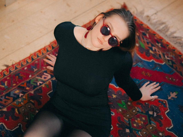 MaryWaddell with dark sunglasses, black dress, and black tights, sitting on a colorful patterned rug, looking up with hands on the rug, showing her tits and ass.
