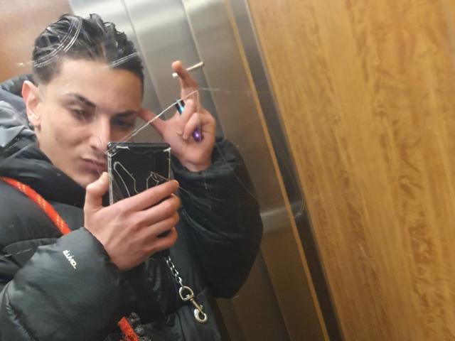 HispanoX with short, dark hair, puffy black jacket, and orange strap, holding phone, smoking, making peace sign, wooden wall reflection, elevator setting.