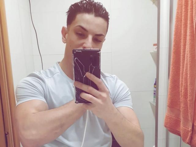 HispanoX with short dark wet hair, muscular arms, white t-shirt, holding black phone with white lines, taking selfie in bathroom, orange towel on right.
