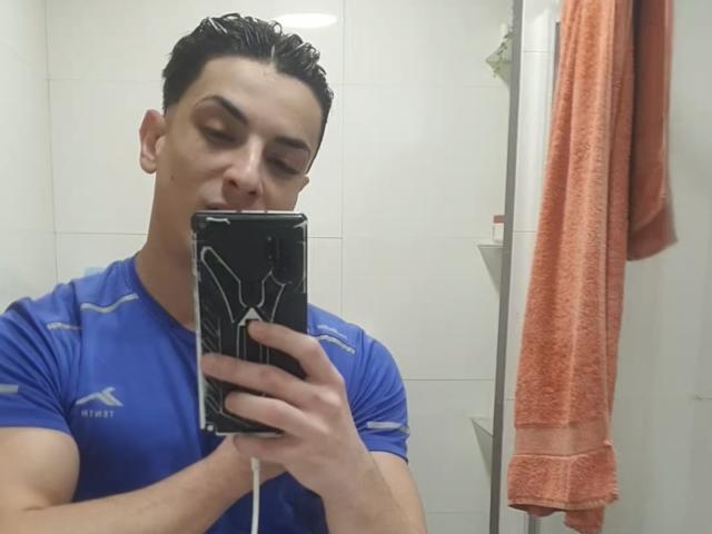 HispanoX with wet, dark hair, wearing a blue sports shirt, takes a mirror selfie with a black phone, showing a white logo. Orange towel hangs to the right.