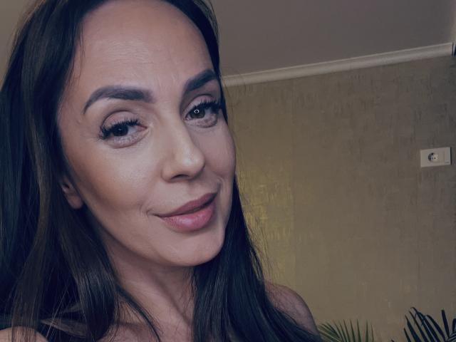 JenniferHills has long brown hair, arched eyebrows, and is smiling with pink lipstick. Her eyes are slightly upturned, and she has a subtle, confident look.