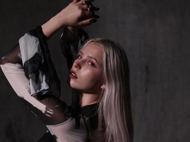 AliceWhitey with long blonde hair, black and white sheer top, arms raised, dark nails, pale skin, and pouty lips, in a moody, dark-lit shot.