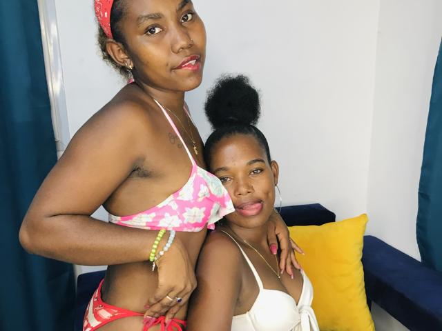 StellaTruda with dark skin wearing a pink floral bikini top and red bottom, standing next to StellaTruda in a white bra. Both have curly hair, and StellaTruda in bikini has a necklace and bracelet.