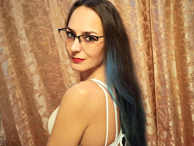 MilfGinna-hot with long blue-tinted hair, red lipstick, black glasses, white bra, smiling over shoulder, looking back. Boobs visible from side. Elegant patterned curtain in background.
