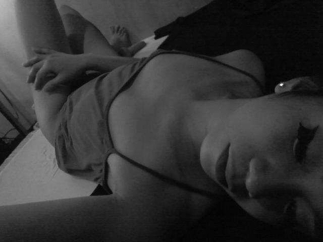Black-and-white photo of xxSexyCumTsDomxx lying on bed, wearing a loose tank top, exposing her small tits and flat stomach, with her hand on her thigh, legs raised.