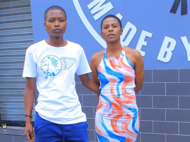 Two sexyboobaex5s standing, one in a white t-shirt with blue and white logo, blue pants, other in a colorful, wavy dress, arms behind back, against a blue wall.