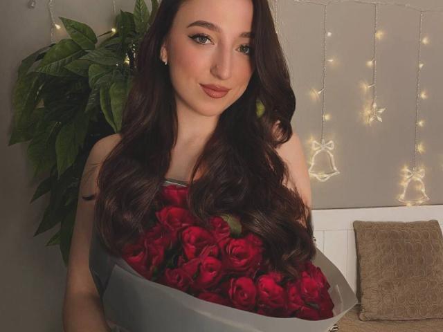 MonAmourR, com cabelos longos e castanho-escuros, segurando um buquê de rosas vermelhas, sorrindo suavemente, usando maquiagem discreta e pequenos brincos, em um quarto com iluminação suave..