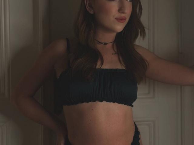 MonAmourR, con i capelli castani e ondulati, indossa un reggiseno a fascia nero con drappeggi, una collana choker e rossetto rosso, è in piedi con le mani sui fianchi, mostrando il suo addome tonico e i suoi piccoli seni..