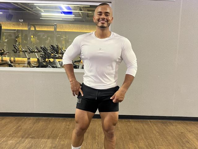 JeronimoHot stands in a gym, wearing a white long-sleeve shirt and black shorts, smiling with hands on hips, showing a muscular, toned body and shaved head.