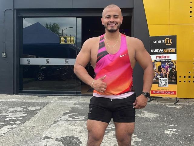 JeronimoHot, bald with a beard, muscular, wearing a pink, orange, and purple gradient tank top and black shorts, giving a thumbs-up, standing in front of a building.