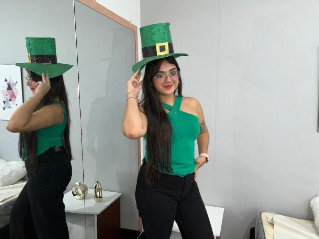 VictoriaSaens with long black hair, green sleeveless top, black pants, and green hat with black band and gold buckle, standing, pointing at hat, mirror reflection, smiling.