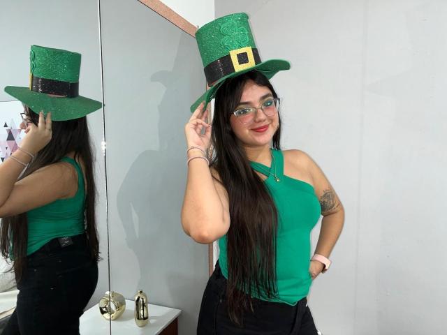 VictoriaSaens with green top hat, glasses, and long black hair, touching hat, green halter top, black pants, tattoo on right arm, reflection in mirror, smiling.