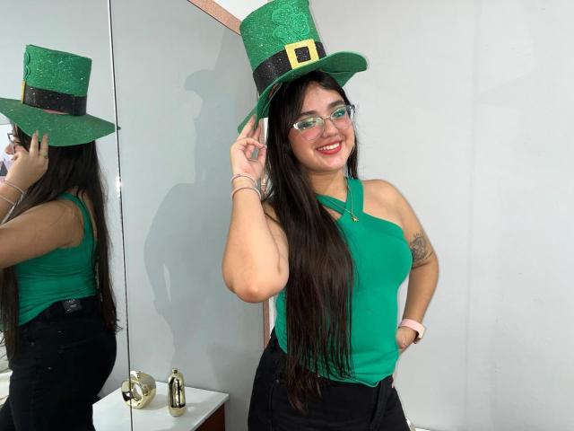 VictoriaSaens with green top hat, green halter top, black skirt, glasses, and red lipstick, showing her ass with hand on hip, smiling, long brown hair, tattoo on arm.