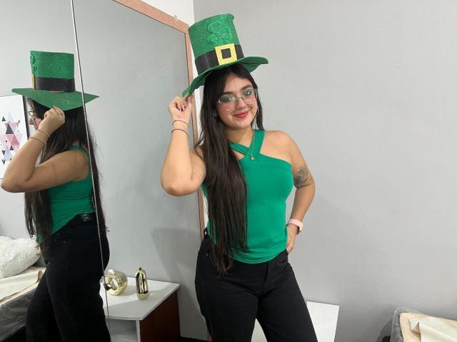 VictoriaSaens with long black hair, green eyes, glasses, green top, black pants, green Leprechaun hat, holding hat, mirror reflection, tattoo on right arm, smiling, pink watch.