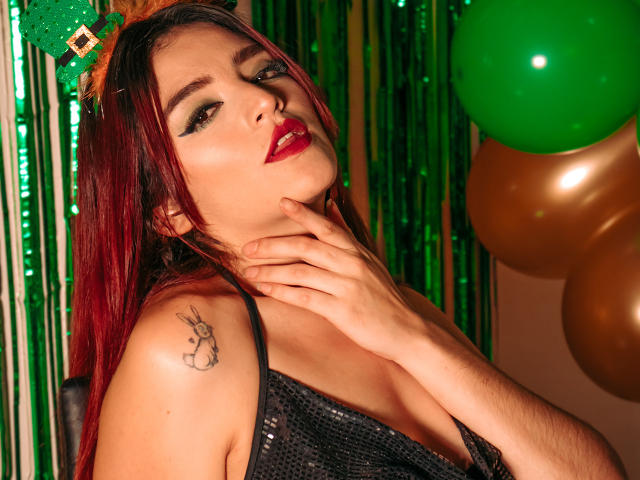 ArianaWithe with red lipstick, black sequined top, green and gold balloons, green streamers, and a tattoo of a bunny on her right shoulder, hand on neck, looking at camera.