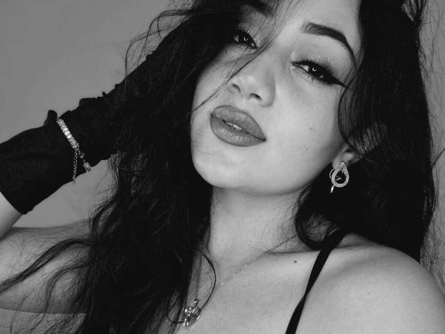 IsaSweets with dark, wavy hair, full lips, and dramatic eyeliner. She's wearing a black glove, earrings, and a necklace, looking seductive with a hand in her hair. Black and white.