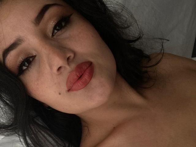 IsaSweets with dark hair, full red lips, and brown eyes, lying down, showing freckles and a small beauty mark on her cheek. She's topless, with visible shoulders.