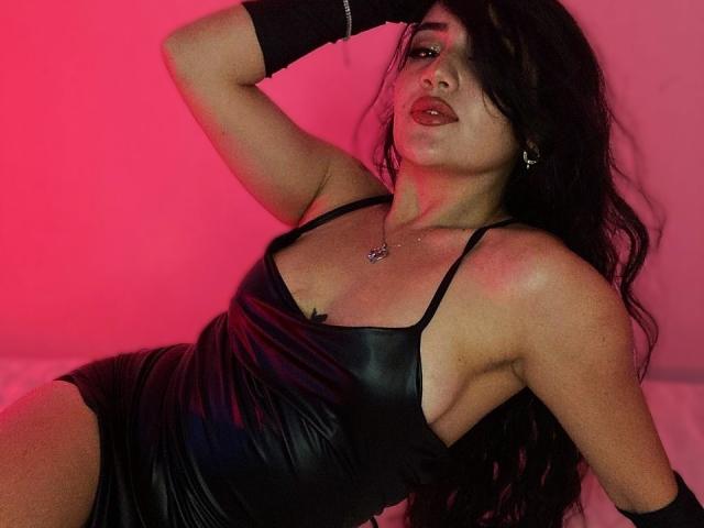 IsaSweets in a black leather dress, showing her tits, with long black hair, red lighting, and a sexy pose, wearing a black glove and necklace.