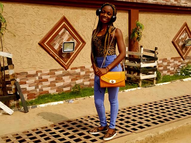 Ruzzyde1-hot with dark skin, wearing headphones, leopard print top, blue jeans, brown sneakers, orange crossbody bag, standing on sidewalk, smiling.