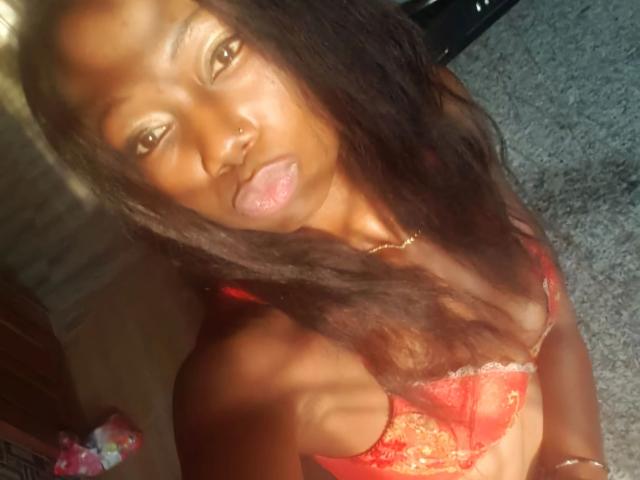 Ruzzyde1-hot with dark skin, long black hair, nose ring, gold necklace, red lace bra, tongue out, standing on grey carpet, sunlight on face, upper body shot.