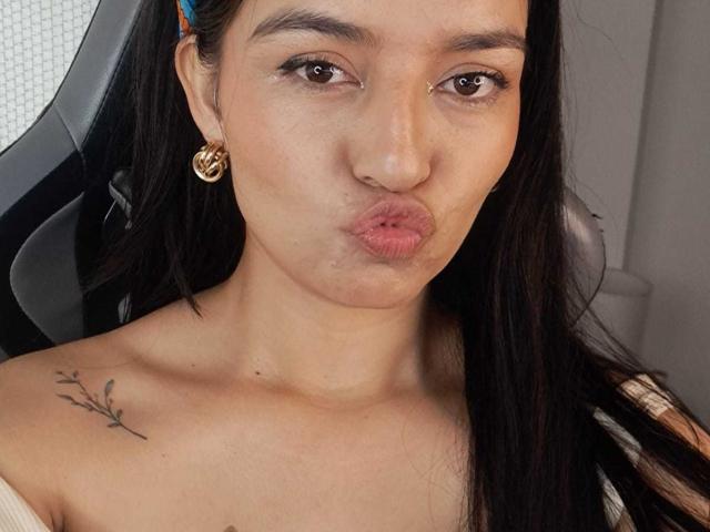 HathorX with pouty lips, brown eyes, long black hair, gold hoop earrings, small tattoo on right shoulder, wearing off-shoulder top, in chair, making kissy face.