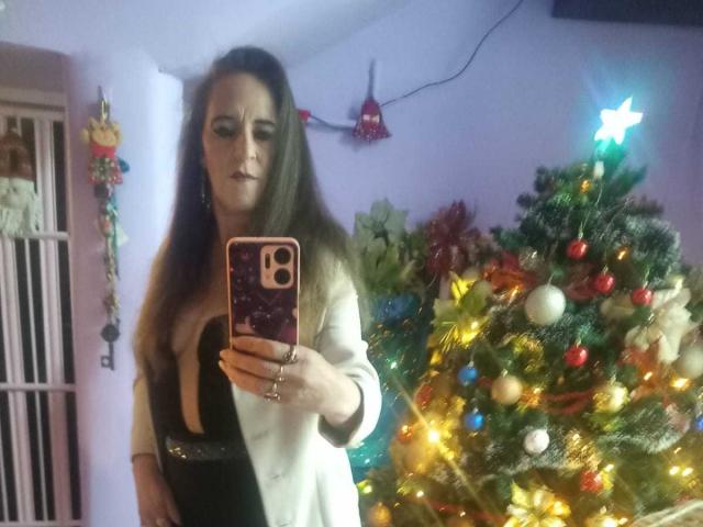 ChanellaMature with long brown hair, white blazer, black dress, holding phone, standing in front of Christmas tree with colorful lights, taking selfie. Boobs visible under dress.