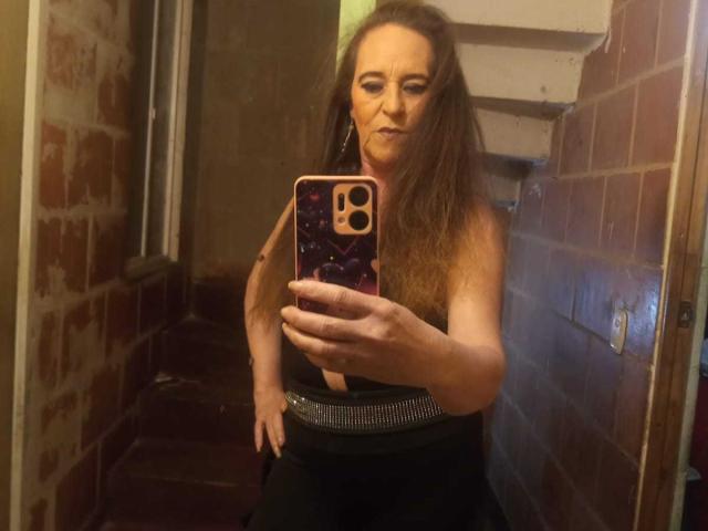 ChanellaMature with long brown hair, wearing a black dress with a silver belt, taking a mirror selfie in a dimly lit, narrow hallway, holding an iPhone with a purple case.