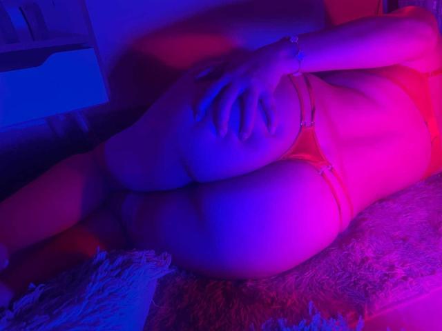 ZoeEmerson lying on a bed, legs bent, wearing pink lingerie, hands on her ass, purple and pink lights, bracelet on her wrist, fuzzy blanket, visible butt, relaxed pose.
