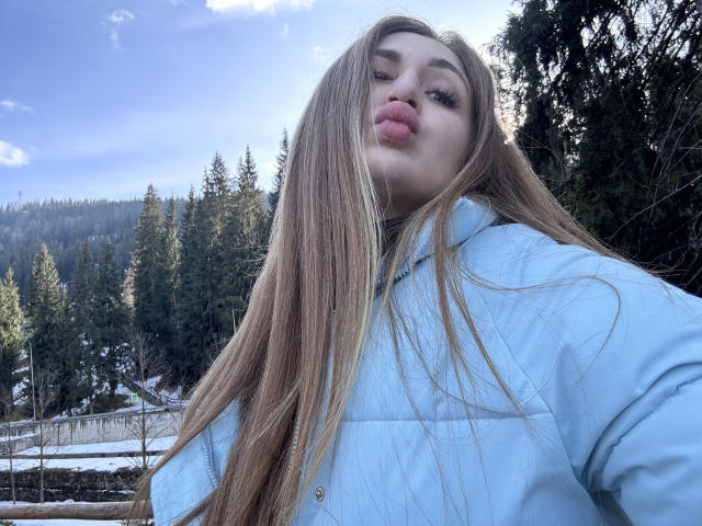 BeataAngel with long blonde hair, puffy blue jacket, and pouty pink lips, looking down at the camera in a snowy forest. Her ass is partially visible.